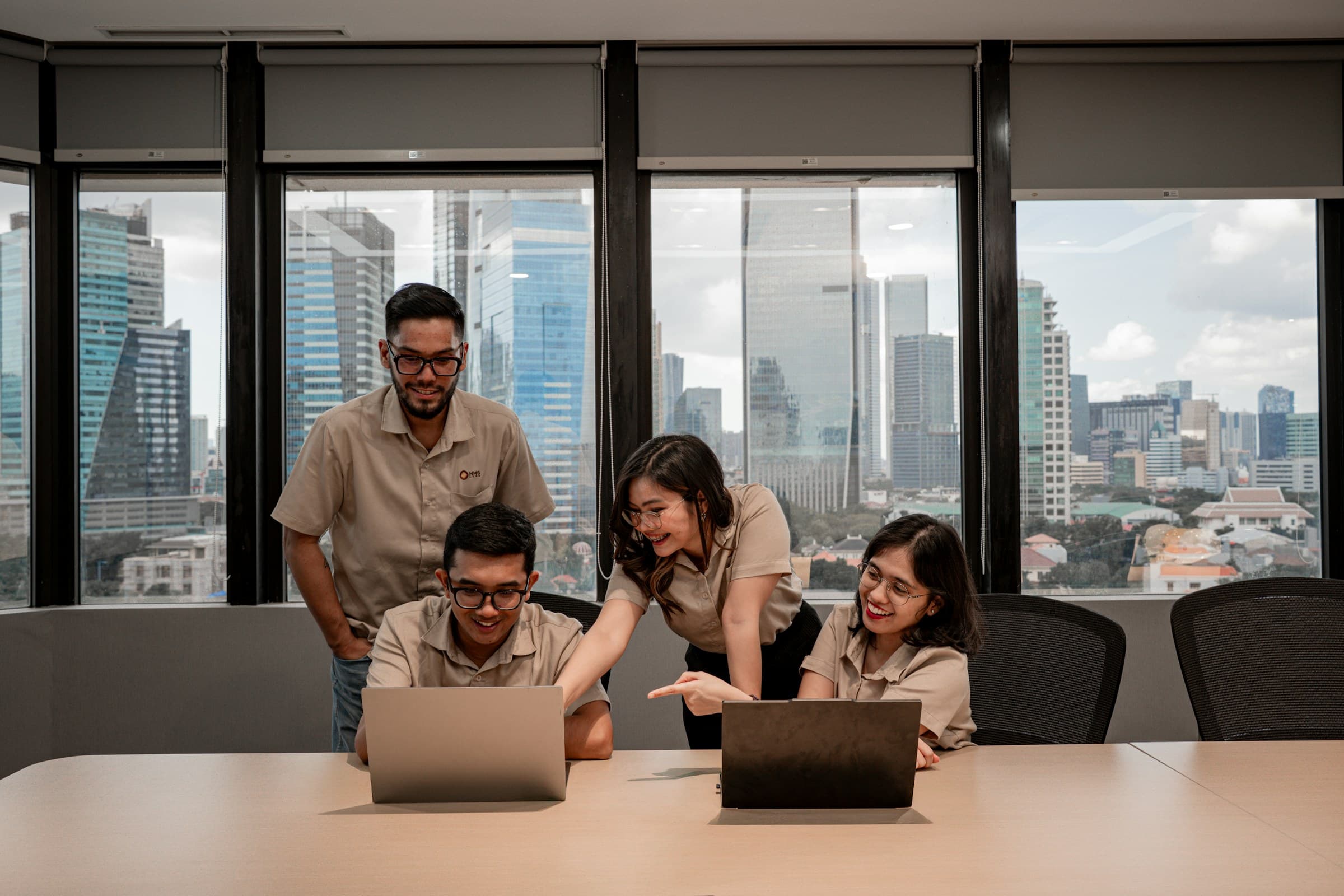 Four colleagues around a board table working from two laptops, a city skyline filling the windows behind them.